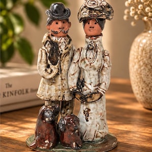May include: A ceramic figurine set featuring a couple in textured white attire with brown accents, standing on a circular base. The figure on the left wears a dark hat and mustache, while the figure on the right wears a wide-brimmed hat. Two small brown dogs sit at their feet.