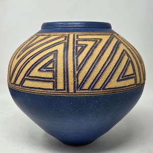 May include: A round ceramic vase with a deep blue matte finish. The upper section features a wide band of textured, tan-colored geometric patterns consisting of nested triangles and parallel lines carved into the surface of the clay vessel.