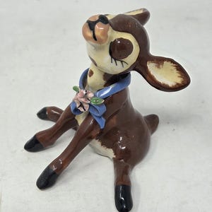 May include: A glossy ceramic figurine of a seated fawn with dark brown fur, cream-colored accents on the face and ears, and black hooves. The fawn wears a blue ribbon collar adorned with a small cluster of pink flowers and green leaves.