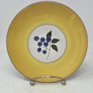 May include: A round ceramic plate featuring a wide, solid yellow border and a white center circle. The center of the plate displays a painted illustration of a cluster of small blue berries on a brown stem with several dark green leaves.