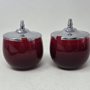 Könnte beinhalten: Zwei passende Aschenbecher in Rot und Chrom. Die Aschenbecher haben einen abgerundeten, eiförmigen Körper mit einer glänzenden, dunkelroten Oberfläche, die an der Basis in Schwarz übergeht. Jeder Aschenbecher hat einen Chromdeckel und einen kleinen, zylindrischen Chromgriff.