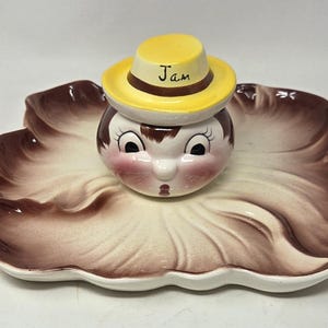 May include: A vintage ceramic jam jar with a whimsical face design, topped with a yellow hat labeled "Jam." The jar sits on a leaf-shaped serving dish with brown and cream tones. A charming and decorative kitchenware item.