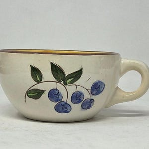 May include: A cream-colored ceramic mug featuring a hand-painted design of blue berries and green leaves on the side. The mug has a rounded bowl shape, a single handle on the right side, and a dark brown rim around the top edge of the vessel.