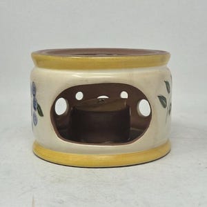 May include: A cylindrical ceramic oil warmer featuring a cream-colored body with yellow rims at the top and bottom. The exterior is decorated with painted blue berries and green leaves. It has large oval cutouts and a dark brown interior base.