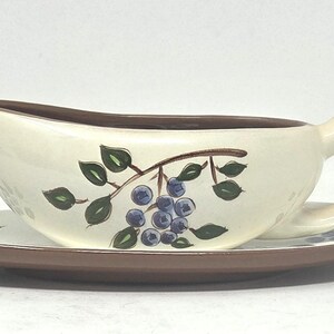 May include: A cream-colored ceramic gravy boat with a brown rim and handle, featuring a hand-painted design of blue berries and green leaves on the side. The gravy boat rests on a matching oval saucer with a brown edge and a berry motif.
