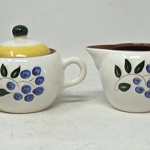 May include: A ceramic sugar bowl with a yellow lid and a matching creamer pitcher. Both pieces are off-white with a hand-painted pattern of blue berries and green leaves. The sugar bowl features two side handles and a dark green knob on the lid.