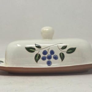 May include: A ceramic butter dish with a cream-colored lid and a terracotta-colored base. The lid features a hand-painted design of blue berries and green leaves on a vine. The lid has a small, rounded knob handle on top for easy lifting.