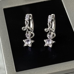 Puede incluir: Un par de pendientes de aro plateados con un dije en forma de estrella. Cada pendiente presenta una pequeña gema transparente y un colgante de estrella con una gema púrpura central. Los pendientes se muestran sobre una superficie negra.