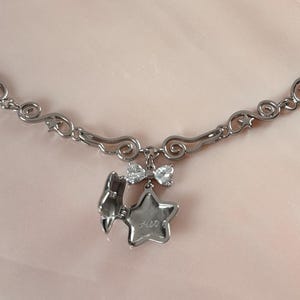 May include: A silver-toned necklace with a decorative chain and a pendant featuring a star and a lightning bolt charm. The necklace has a delicate design with a lobster clasp closure. The pendant also includes a small bow and a small gem.