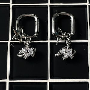 May include: A pair of silver earrings with a dangling orb charm and a star charm. The orb charm is decorated with clear rhinestones.