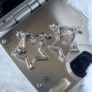 May include: Two silver star-shaped earrings with pink crystal accents and a small arrow charm hanging from each. The earrings are on a silver camera.