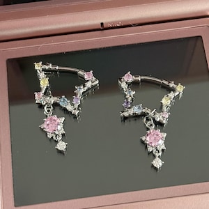 Könnte beinhalten: Ein Paar silberfarbene Ohrringe mit sternförmigem Design. Jeder Ohrring hat einen gebogenen Stab mit bunten Edelsteinen und einen hängenden rosa Blumenanhänger mit einem kleinen diamantartigen Anhänger. Die Ohrringe werden auf einem rosa Handheld-Spielgerät präsentiert.