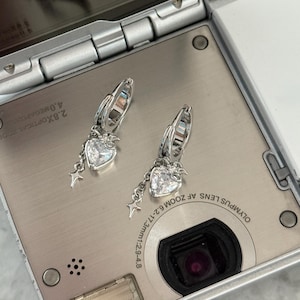 May include: Two silver hoop earrings with heart-shaped clear gemstones and dangling star charms. The earrings are displayed on a silver digital camera.