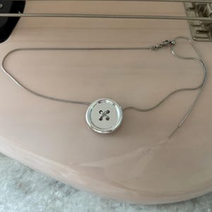Peut inclure: Un collier en argent avec un grand pendentif rond en forme de bouton avec un motif de croix. Le collier repose sur le corps d'une basse rose, avec les cordes et le matériel visibles. Le fond est une surface claire et texturée.