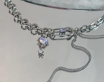 Op de afbeelding: Een zilverkleurige ketting met een kettingontwerp. De ketting heeft een veiligheidsspeldsluiting met een paarse edelsteen en een bungelende bedel met een hartvormige iriserende steen. Een delicate ketting hangt aan de sluiting.