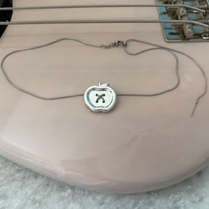Puede incluir: Un collar de plata con un colgante en forma de manzana con un diseño cosido. El collar se muestra en un bajo rosa claro.