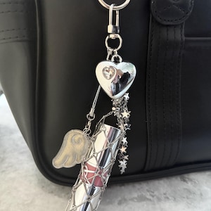 Op de afbeelding: Een zilveren lippenstiftbedel met een roze lippenstiftbuis, een zilveren hart, een gouden vleugel en sterbedels die aan een zilveren ketting hangen. De bedel is bevestigd aan een zwarte tas met zilveren hardware.