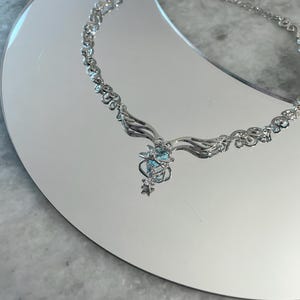 Puede incluir: Un collar de plata con una cadena delicada y un colgante con un racimo de cristales azules y detalles en forma de estrella.