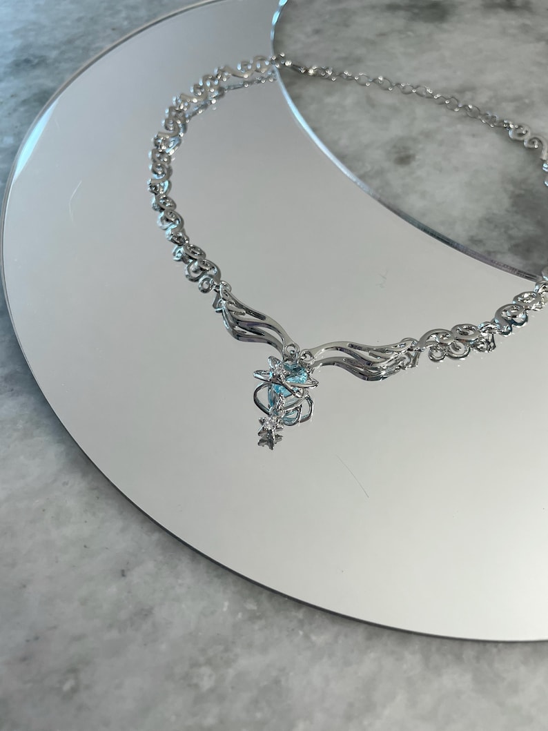 Puede incluir: Un collar de plata con una cadena delicada y un colgante con un racimo de cristales azules y detalles en forma de estrella.