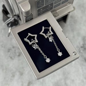May include: Silver star-shaped drop earrings with clear gemstone detailing. The earrings have a star within a star design, with a chain holding a larger gemstone. The earrings are presented on a dark blue surface.