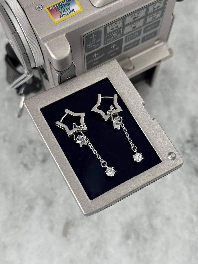 May include: Silver star-shaped drop earrings with clear gemstone detailing. The earrings have a star within a star design, with a chain holding a larger gemstone. The earrings are presented on a dark blue surface.