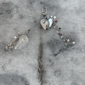 Op de afbeelding: Een zilverkleurige sleutelhanger met een hartvormige bedel, een doorzichtige vleugelbedel en steraccenten. De hartbedel heeft een kleine edelsteen in het midden. Een ketting met een decoratief patroon hangt aan het hart.