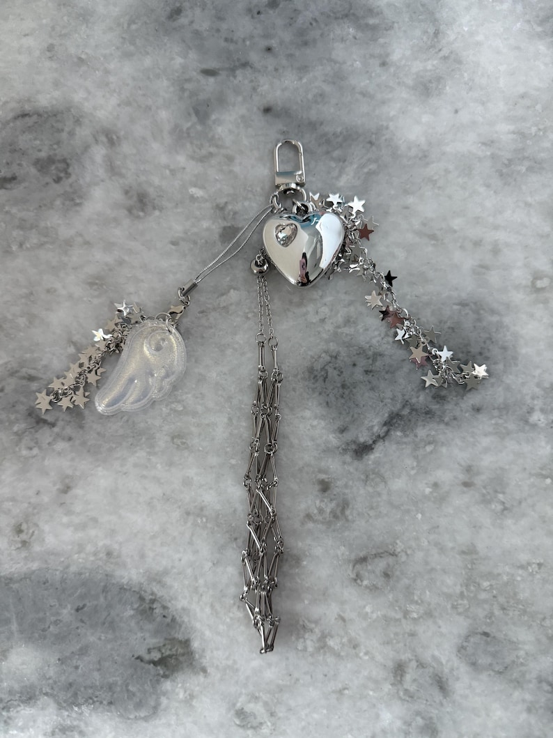 Op de afbeelding: Een zilverkleurige sleutelhanger met een hartvormige bedel, een doorzichtige vleugelbedel en steraccenten. De hartbedel heeft een kleine edelsteen in het midden. Een ketting met een decoratief patroon hangt aan het hart.