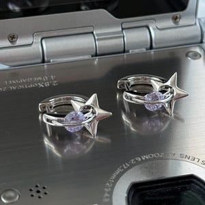 May include: A pair of silver hoop earrings. Each earring features a star-shaped charm and a small, round, light purple gemstone. The earrings are displayed on a silver surface.