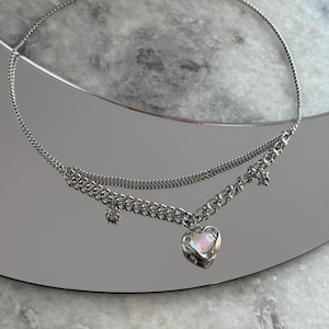 Pode incluir: Um colar prateado com um design em camadas. O colar apresenta uma corrente fina e uma corrente mais grossa com um pingente em forma de cora&ccedil;&atilde;o. Pequenos pingentes de estrela e redondos pendem da corrente. O colar &eacute; exibido em uma superf&iacute;cie reflexiva.