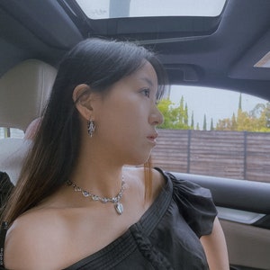 Puede incluir: Una mujer con el pelo largo y negro lleva una blusa negra de hombros descubiertos y un collar plateado con un colgante en forma de coraz&oacute;n. Est&aacute; sentada en un coche con el techo solar abierto.