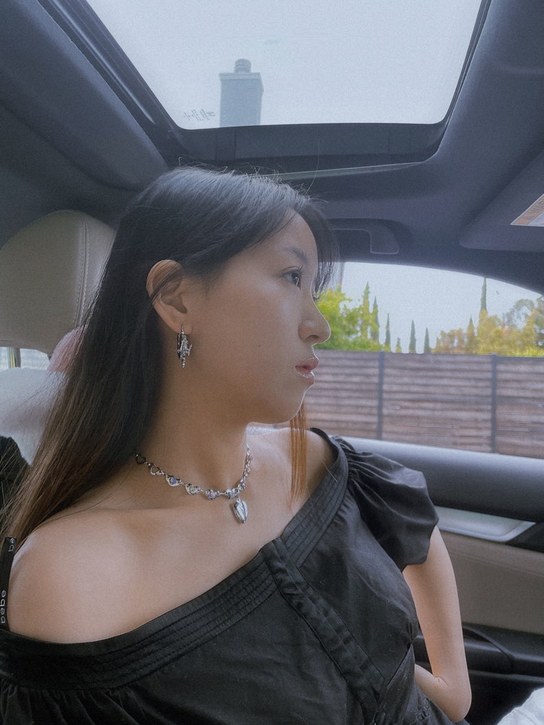 Puede incluir: Una mujer con el pelo largo y negro lleva una blusa negra de hombros descubiertos y un collar plateado con un colgante en forma de coraz&oacute;n. Est&aacute; sentada en un coche con el techo solar abierto.