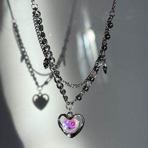 Pode incluir: Um colar prateado em camadas com um pingente de cora&ccedil;&atilde;o. O pingente apresenta uma moldura em forma de cora&ccedil;&atilde;o com um centro iridescente multicolorido. O colar tem uma corrente e um design de contas, com um pequeno pingente pendurado na corrente.