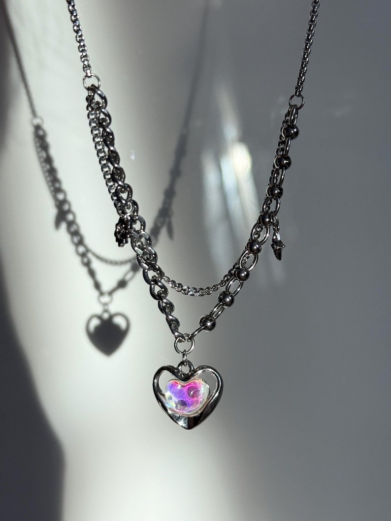 Pode incluir: Um colar prateado em camadas com um pingente de cora&ccedil;&atilde;o. O pingente apresenta uma moldura em forma de cora&ccedil;&atilde;o com um centro iridescente multicolorido. O colar tem uma corrente e um design de contas, com um pequeno pingente pendurado na corrente.