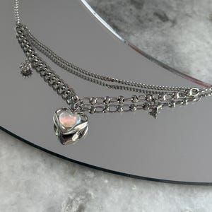 Pode incluir: Um colar de prata em camadas com um pingente de cora&ccedil;&atilde;o. O pingente de cora&ccedil;&atilde;o tem uma pedra iridescente. O colar tem uma corrente e uma corrente delicada com um pequeno pingente de estrela e flor. O colar &eacute; exibido em uma superf&iacute;cie reflexiva.