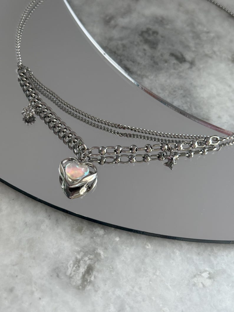 Pode incluir: Um colar de prata em camadas com um pingente de cora&ccedil;&atilde;o. O pingente de cora&ccedil;&atilde;o tem uma pedra iridescente. O colar tem uma corrente e uma corrente delicada com um pequeno pingente de estrela e flor. O colar &eacute; exibido em uma superf&iacute;cie reflexiva.