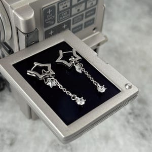 May include: Silver star-shaped drop earrings with a small dangling clear gem. The earrings are displayed on a black screen, with a silver video camera in the background. The earrings have a delicate chain.