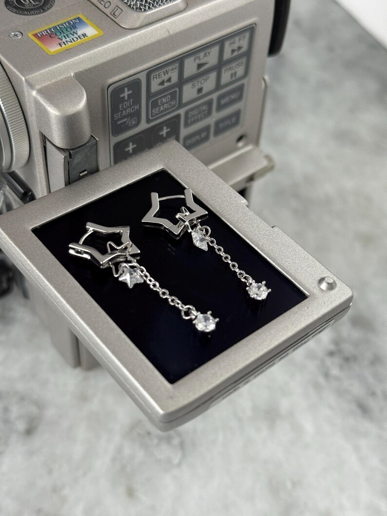 May include: Silver star-shaped drop earrings with a small dangling clear gem. The earrings are displayed on a black screen, with a silver video camera in the background. The earrings have a delicate chain.