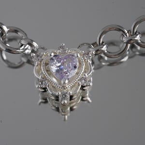 May include: A silver necklace with a heart-shaped pendant. The pendant features a light purple gemstone at its center, surrounded by smaller clear stones and ornate silver detailing. The chain is made of large, circular links.