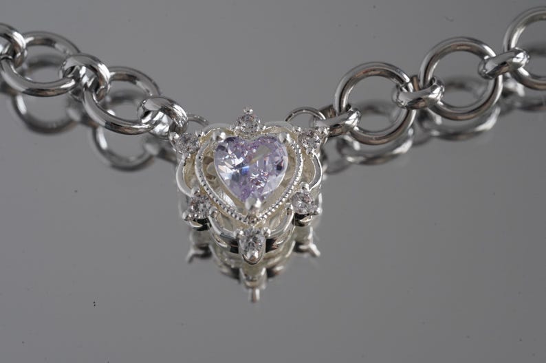May include: A silver necklace with a heart-shaped pendant. The pendant features a light purple gemstone at its center, surrounded by smaller clear stones and ornate silver detailing. The chain is made of large, circular links.