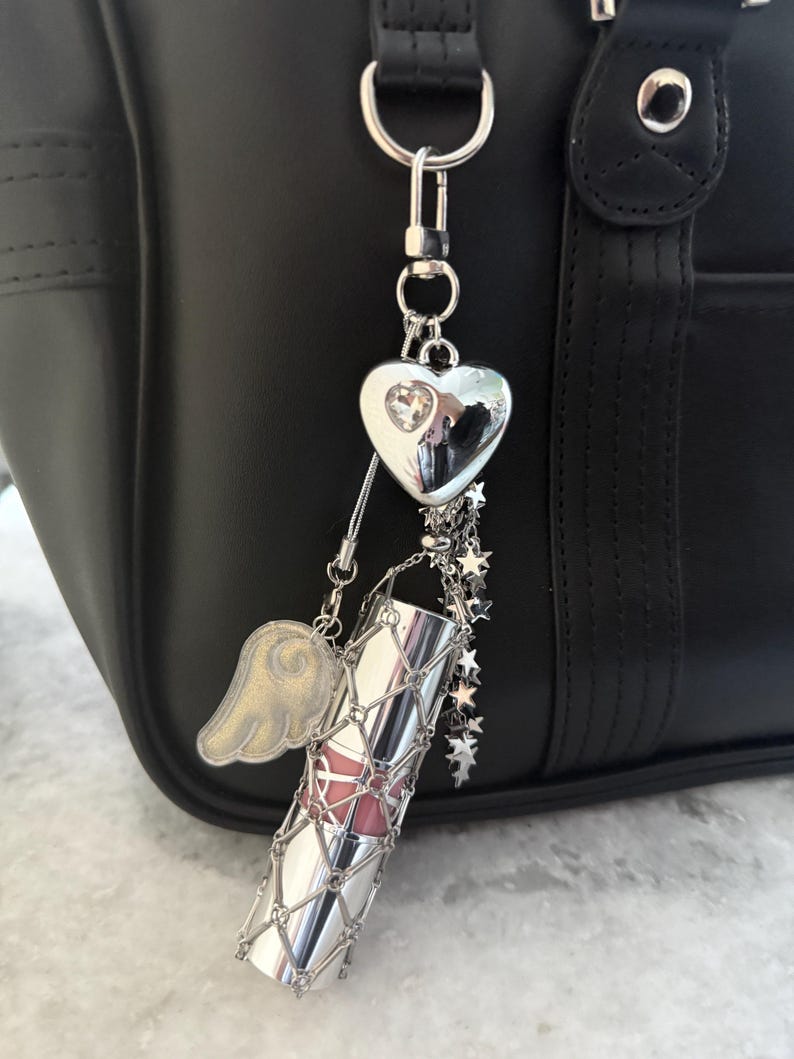 Op de afbeelding: Een zilveren lippenstiftbedel met een roze lippenstiftbuis, een zilveren hart, een gouden vleugel en sterbedels die aan een zilveren ketting hangen. De bedel is bevestigd aan een zwarte tas met zilveren hardware.