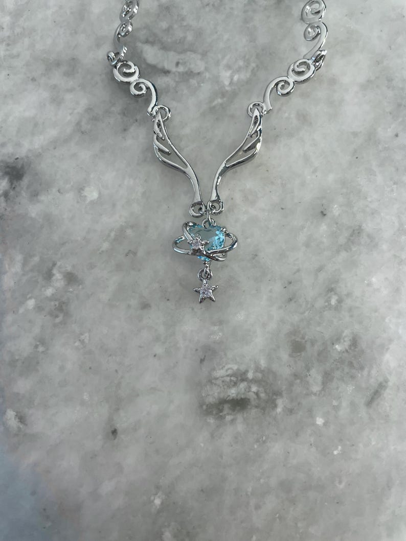 Puede incluir: Collar de plata con un delicado dise&ntilde;o de alas y un colgante de cristal azul. El colgante presenta un peque&ntilde;o amuleto de estrella.