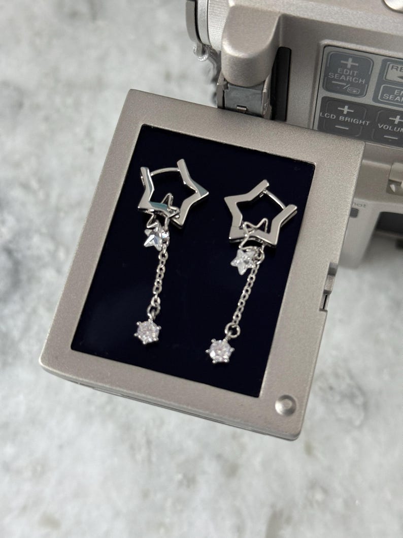 May include: A pair of silver star-shaped earrings. Each earring has a star-shaped hoop, a dangling chain, and a sparkling, round gemstone. The earrings are presented on a dark blue surface.