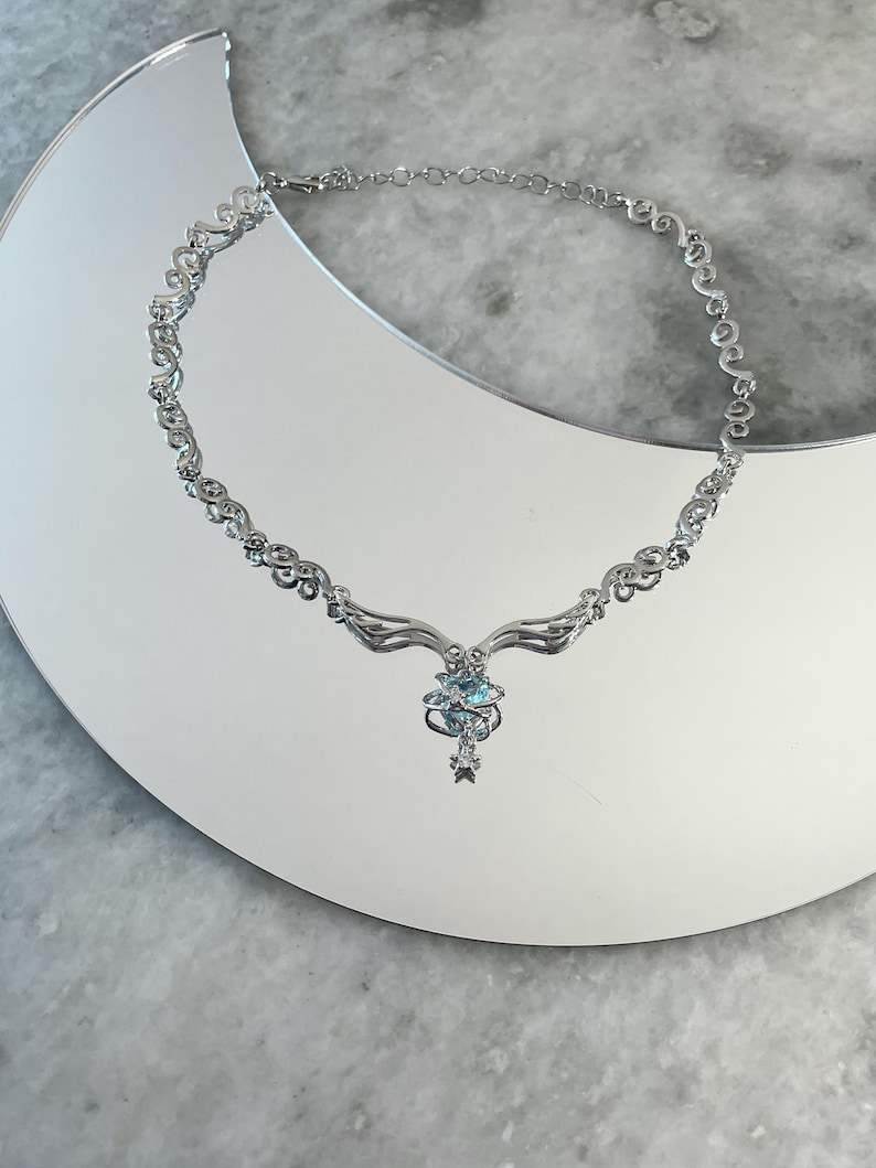 Puede incluir: Un collar de plata con un dise&ntilde;o delicado e intrincado. El colgante presenta un grupo de piedras preciosas azules y una estrella.