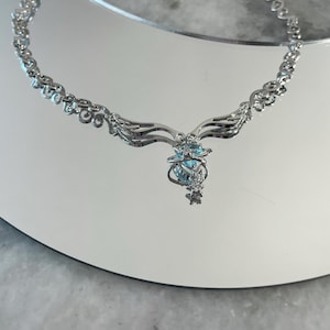 Puede incluir: Un collar de plata con una cadena delicada y un colgante con una piedra preciosa azul rodeada de piedras m&aacute;s peque&ntilde;as. El colgante tiene forma de alas.