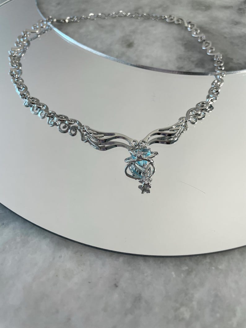 Puede incluir: Un collar de plata con una cadena delicada y un colgante con una piedra preciosa azul rodeada de piedras m&aacute;s peque&ntilde;as. El colgante tiene forma de alas.