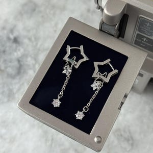 May include: Silver-coloured dangle earrings. Each earring has a star-shaped stud, a smaller star charm, and a chain with a round, clear gemstone. The earrings are presented against a dark blue backdrop, showcasing their celestial design.