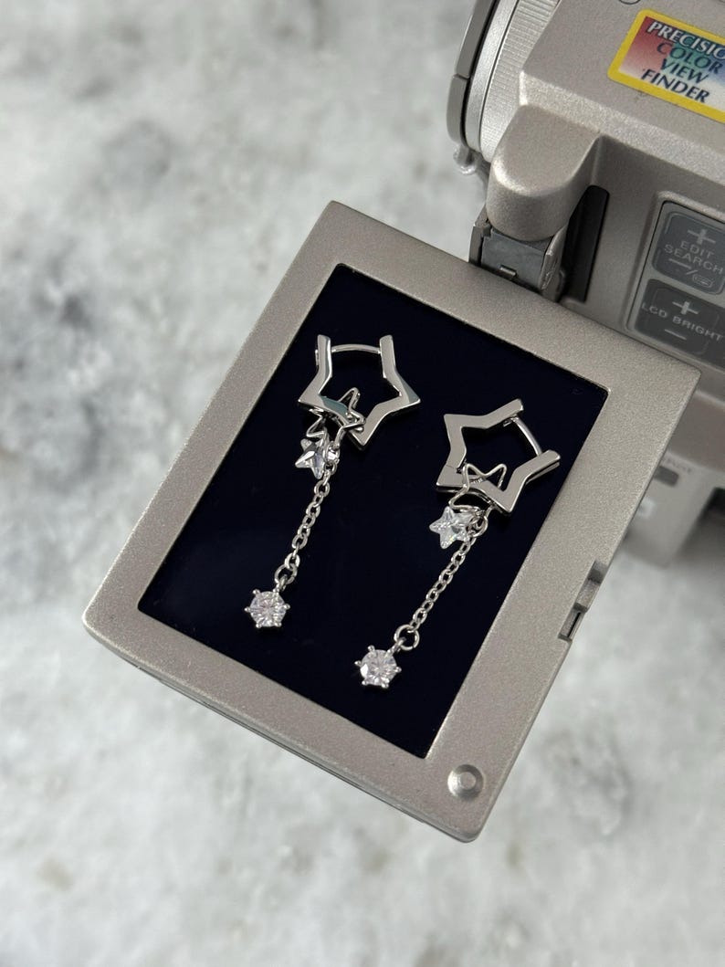 May include: Silver-coloured dangle earrings. Each earring has a star-shaped stud, a smaller star charm, and a chain with a round, clear gemstone. The earrings are presented against a dark blue backdrop, showcasing their celestial design.