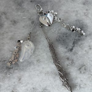 Op de afbeelding: Een zilverkleurige sleutelhanger met een hartvormige bedel met een kleine edelsteen, een doorzichtige vleugelbedel en steraccenten. De sleutelhanger heeft een ketting en een sluiting.