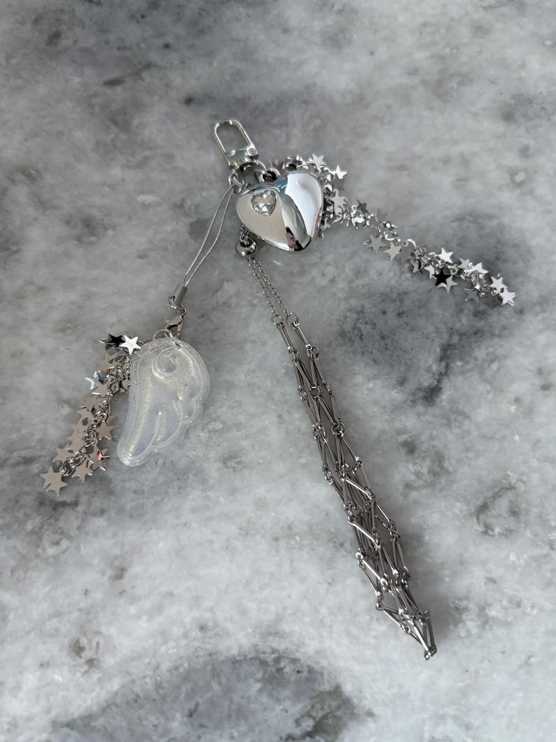 Op de afbeelding: Een zilverkleurige sleutelhanger met een hartvormige bedel met een kleine edelsteen, een doorzichtige vleugelbedel en steraccenten. De sleutelhanger heeft een ketting en een sluiting.