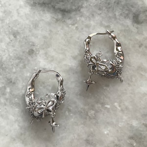 Puede incluir: Un par de pendientes de aro de plata con un dije colgante que presenta un lazo y una estrella. Los pendientes est&aacute;n hechos de metal texturizado y tienen un dise&ntilde;o &uacute;nico y atrevido.